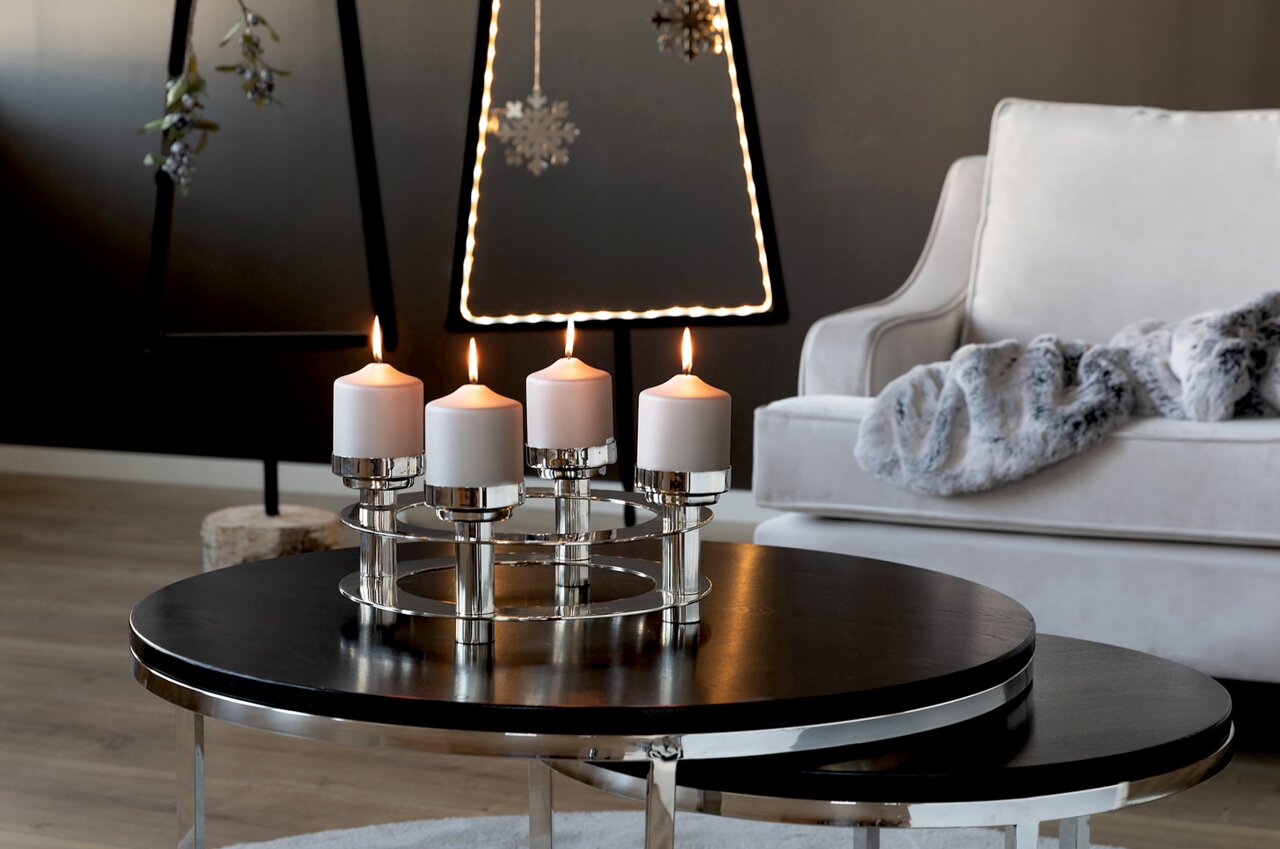 Moderne vierarmige kandelaar van metaal met witte kaarsen, op een ronde tafel in een stijlvol ingerichte woonkamer, gezien vanuit de zijkant.