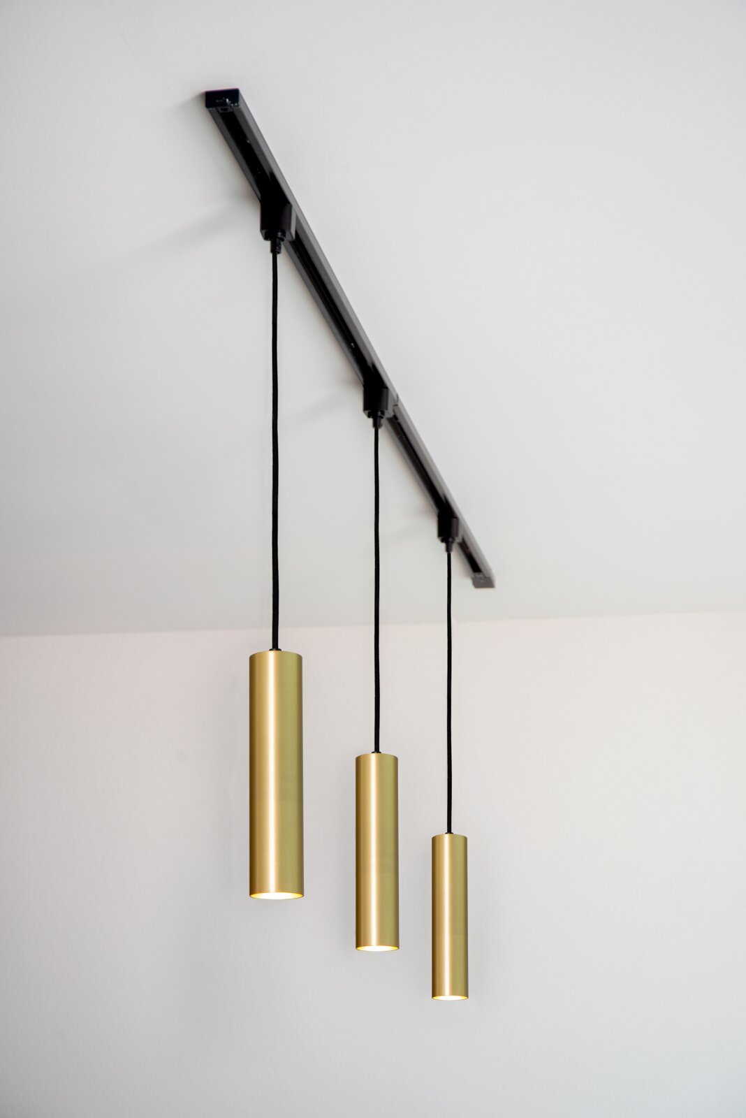 Drei zylindrische Pendelleuchten in mattgoldener Farbe, von einer schwarzen Schiene an der Decke hängend, aus seitlicher Perspektive fotografiert.