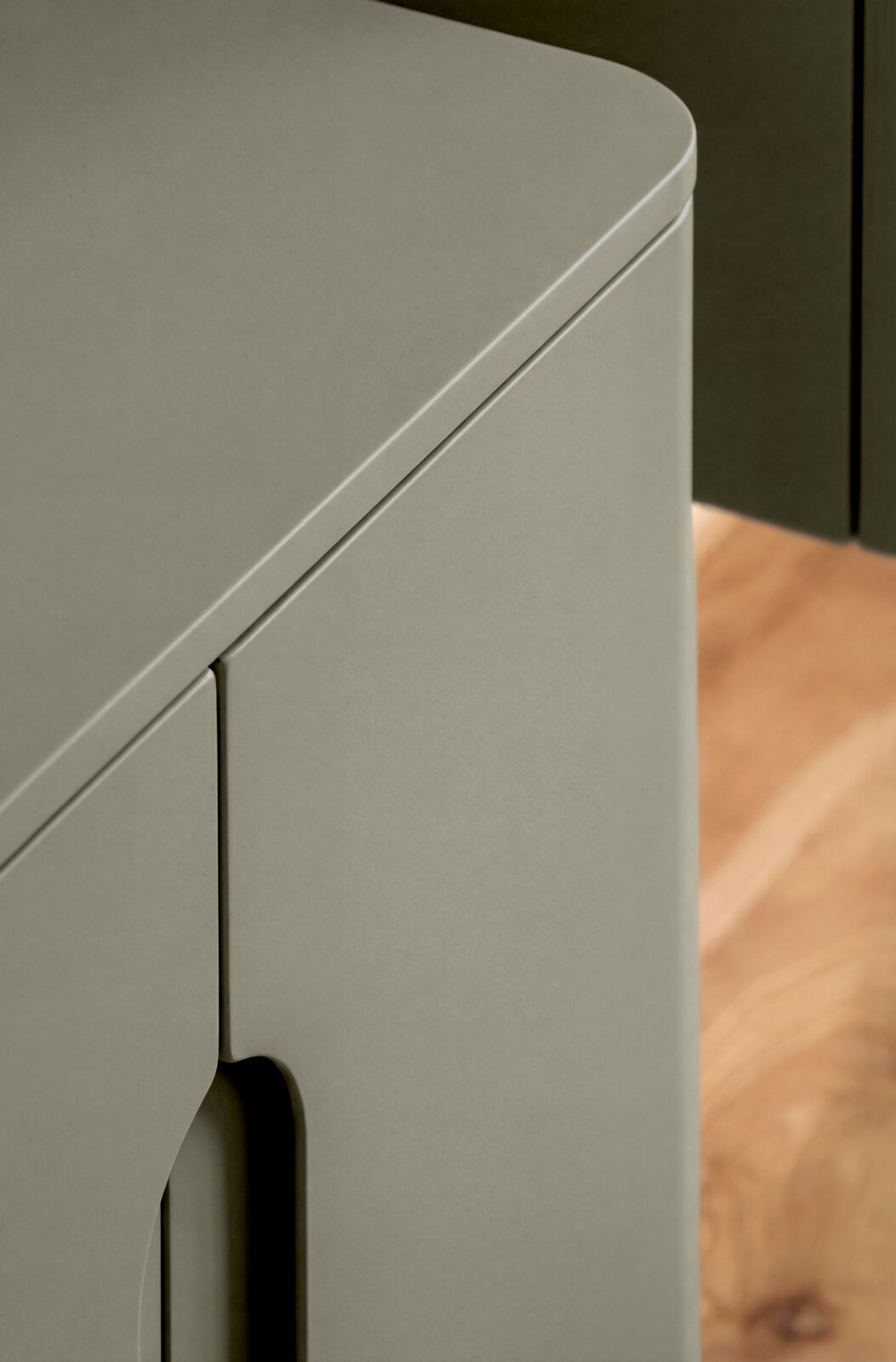 Nahaufnahme eines matten, grauen Schranks/Sideboards mit abgerundeter Oberkante und Türfuge, schräg von oben aus der Ecke fotografiert; Holzboden im Hintergrund.