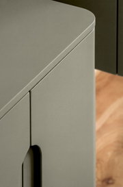 Nahaufnahme eines matten, grauen Schranks/Sideboards mit abgerundeter Oberkante und Türfuge, schräg von oben aus der Ecke fotografiert; Holzboden im Hintergrund.