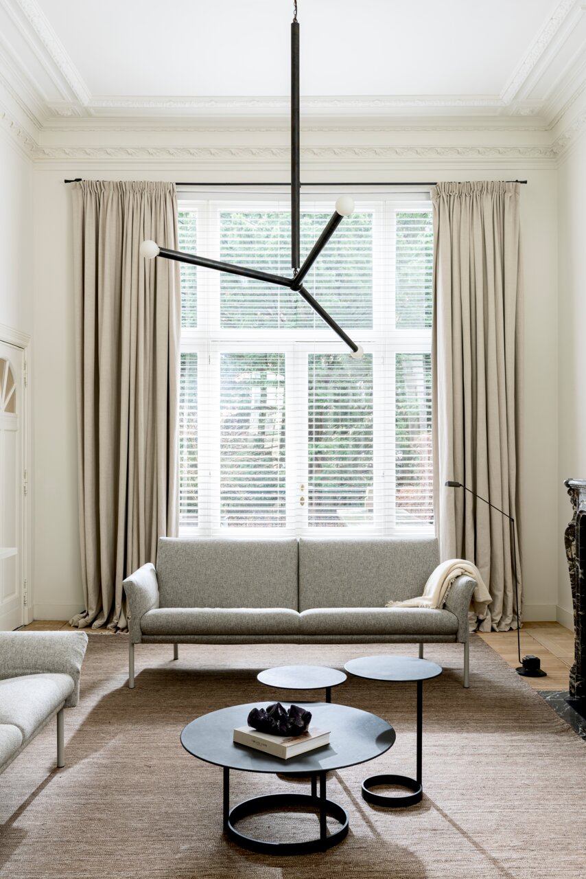 Moderner Wohnraum mit drei runden Beistelltischen in verschiedenen Höhen, zentral im Bild platziert. Im Hintergrund ein großes Fenster mit Vorhängen und ein graues Sofa. Perspektive von vorne.