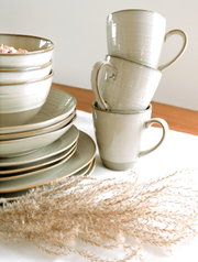 Stapel aus glasierten Keramikbechern in Creme/Beige mit braunem Rand neben passenden Tellern und Schalen auf hellem Tuch, fotografiert in leicht schräger Seitenansicht von vorne