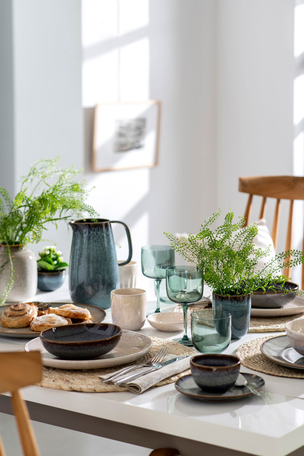 Ein stilvoll gedeckter Tisch mit einem Set aus zwei grünen Weinkelchen im Vordergrund, umgeben von Geschirr, Besteck und Pflanzen in einem hellen Raum.