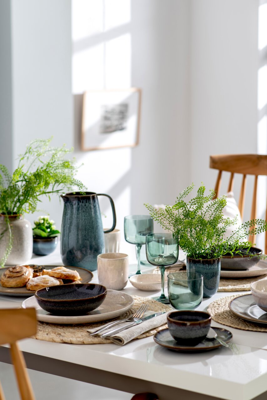 Een stijlvol gedekte tafel met op de voorgrond een set van twee groene wijnglaasjes, omringd door servies, bestek en planten in een lichte ruimte.