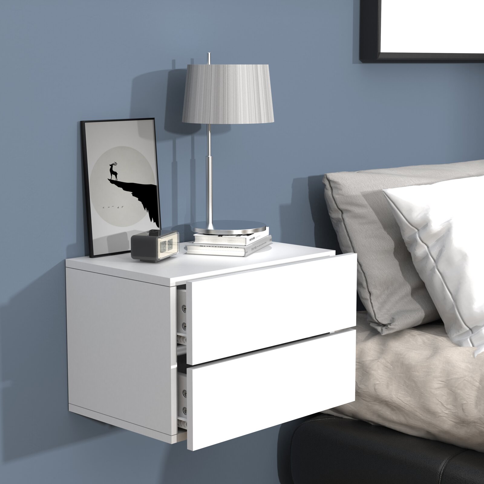 Moderne weiße, wandmontierte Nachttischkommode mit zwei Schubladen neben einem Bett, darauf Tischlampe, Bilderrahmen, Bücher und Wecker Perspektive: frontale leicht schräge Ansicht von links.