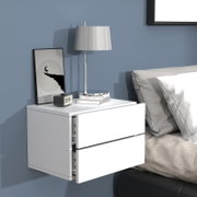 Moderne weiße, wandmontierte Nachttischkommode mit zwei Schubladen neben einem Bett, darauf Tischlampe, Bilderrahmen, Bücher und Wecker; Perspektive: frontale leicht schräge Ansicht von links.
