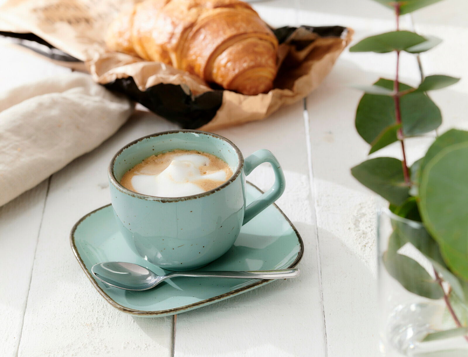Ritzenhoff & Breker Espressountertasse CASA Espressountertasse Casa blau mit Tasse und Löffel, von schräg oben fotografiert, auf einem weißen Holztisch neben einem Croissant und grünen Blättern.