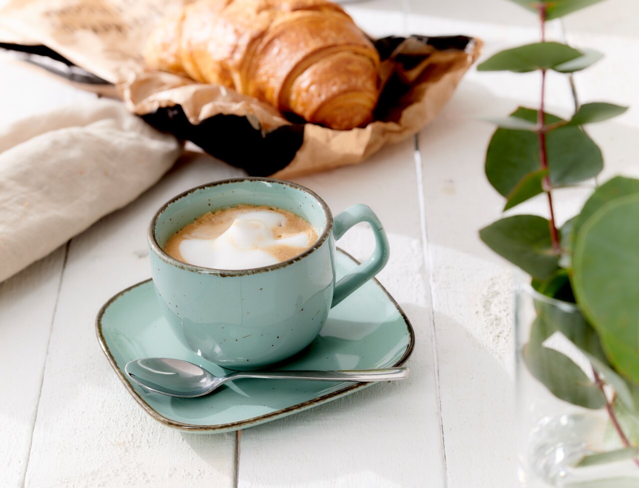 Espressountertasse Casa blau mit Tasse und Löffel, von schräg oben fotografiert, auf einem weißen Holztisch neben einem Croissant und grünen Blättern.