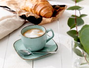Espressountertasse Casa blau mit Tasse und Löffel, von schräg oben fotografiert, auf einem weißen Holztisch neben einem Croissant und grünen Blättern.