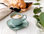 Espressountertasse Casa blau mit Tasse und Löffel, von schräg oben fotografiert, auf einem weißen Holztisch neben einem Croissant und grünen Blättern.