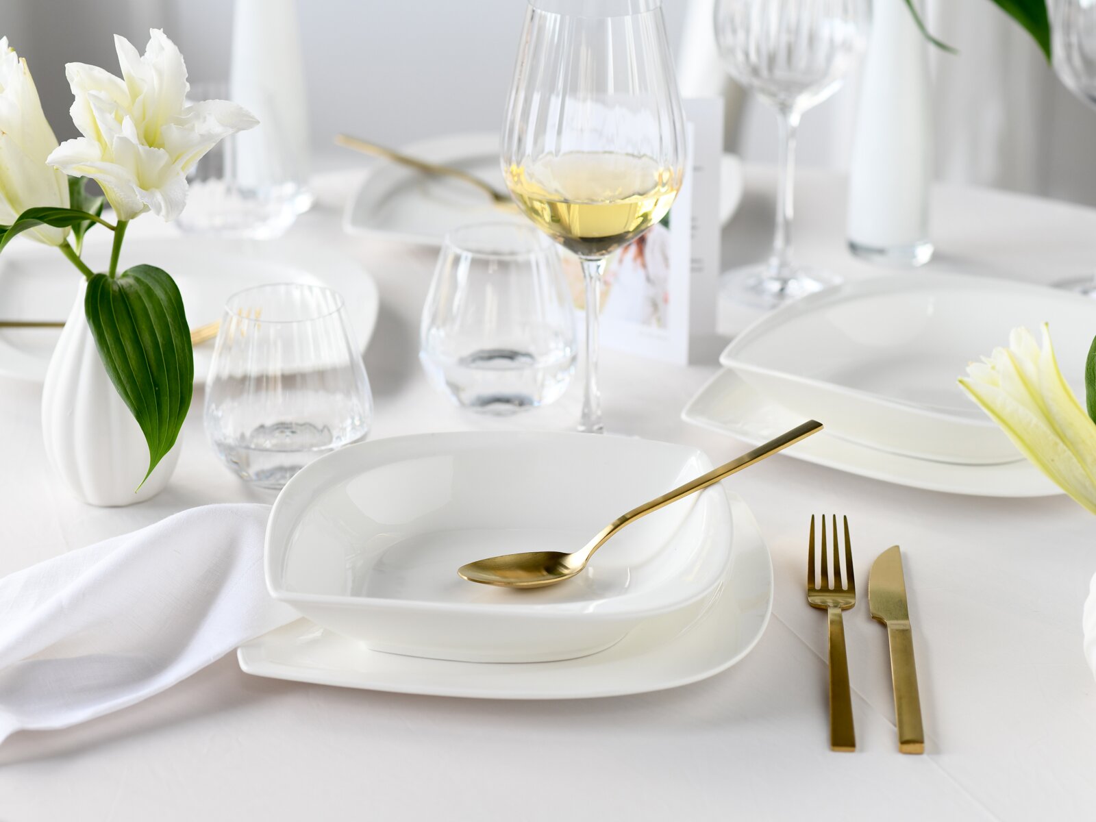 Elegant gedeckter Tisch mit weißem Porzellangeschirr, goldfarbenem Besteck, Wassergläsern und Weißweinglas sowie weißer Blume in Vase, aufgenommen aus leicht schräger Seitenansicht auf Tischhöhe
