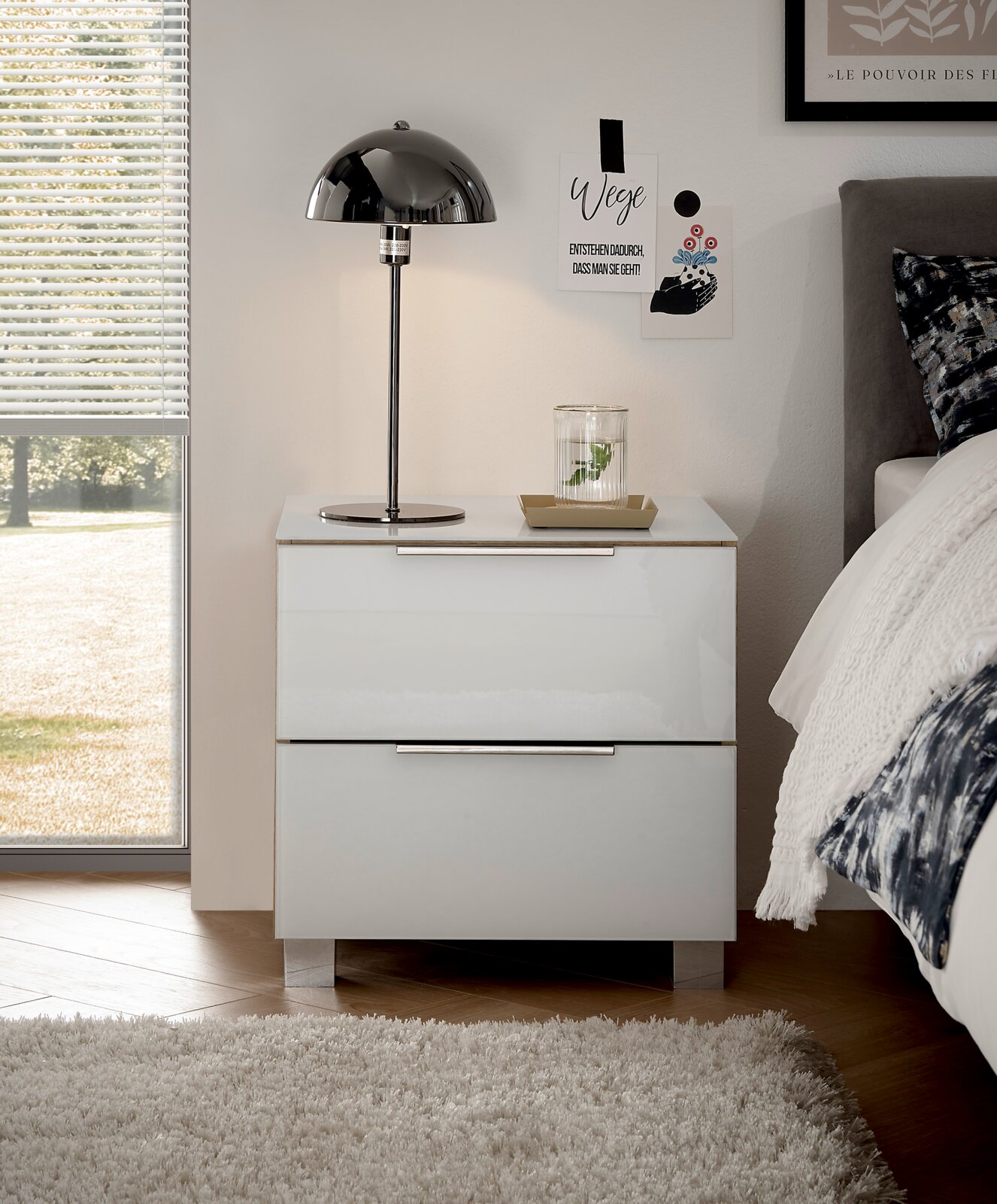 Moderne weiße Nachtkommode mit zwei Schubladen, frontal fotografiert, neben einem Bett mit einer Tischlampe und Dekoration darauf.