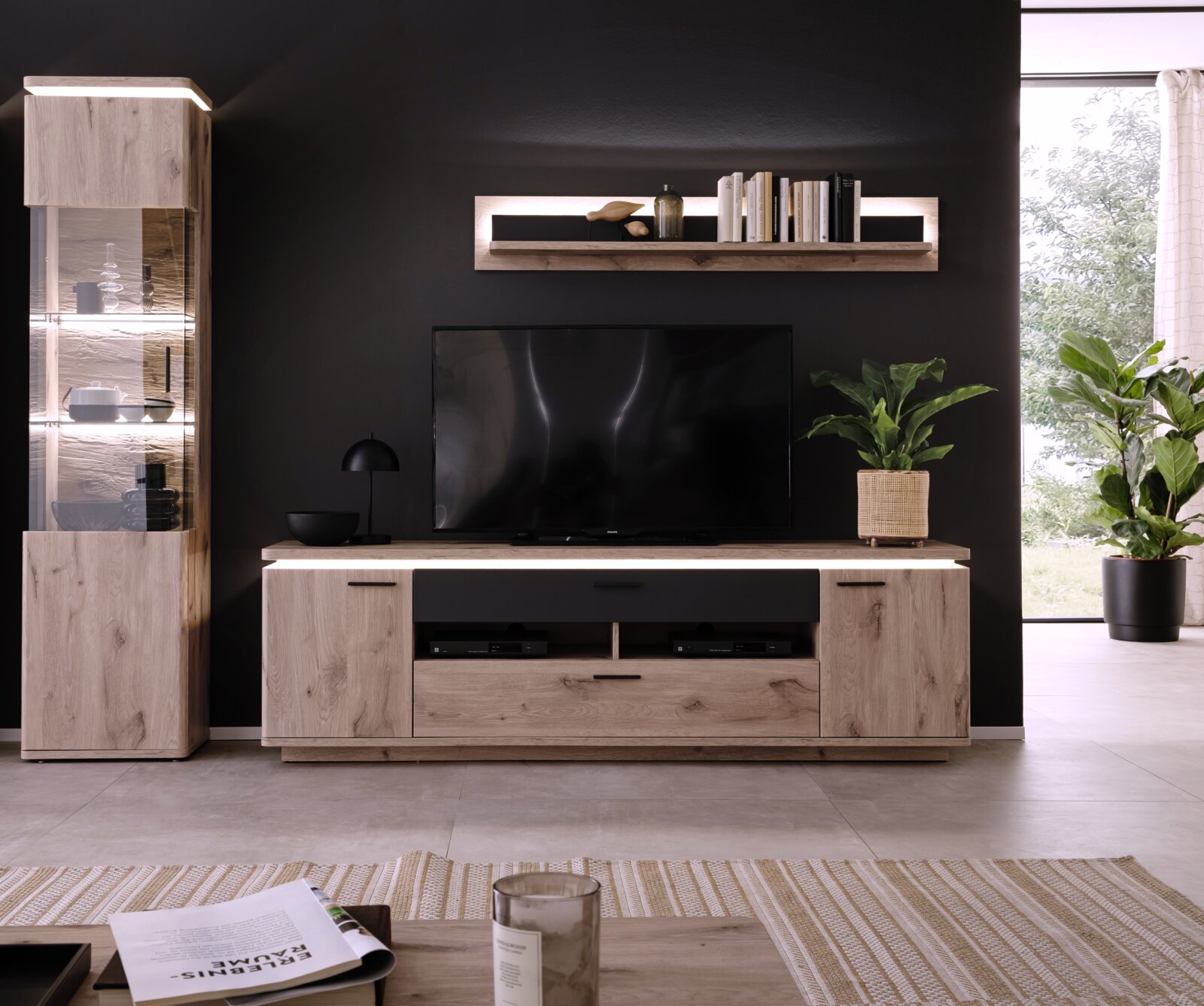 Moderne entertainmentset voor de woonkamer van licht hout, bestaande uit een tv-lowboard, een hoge vitrinekast met glazen deuren en ledverlichting, en een wandplank gefotografeerd vanaf ooghoogte