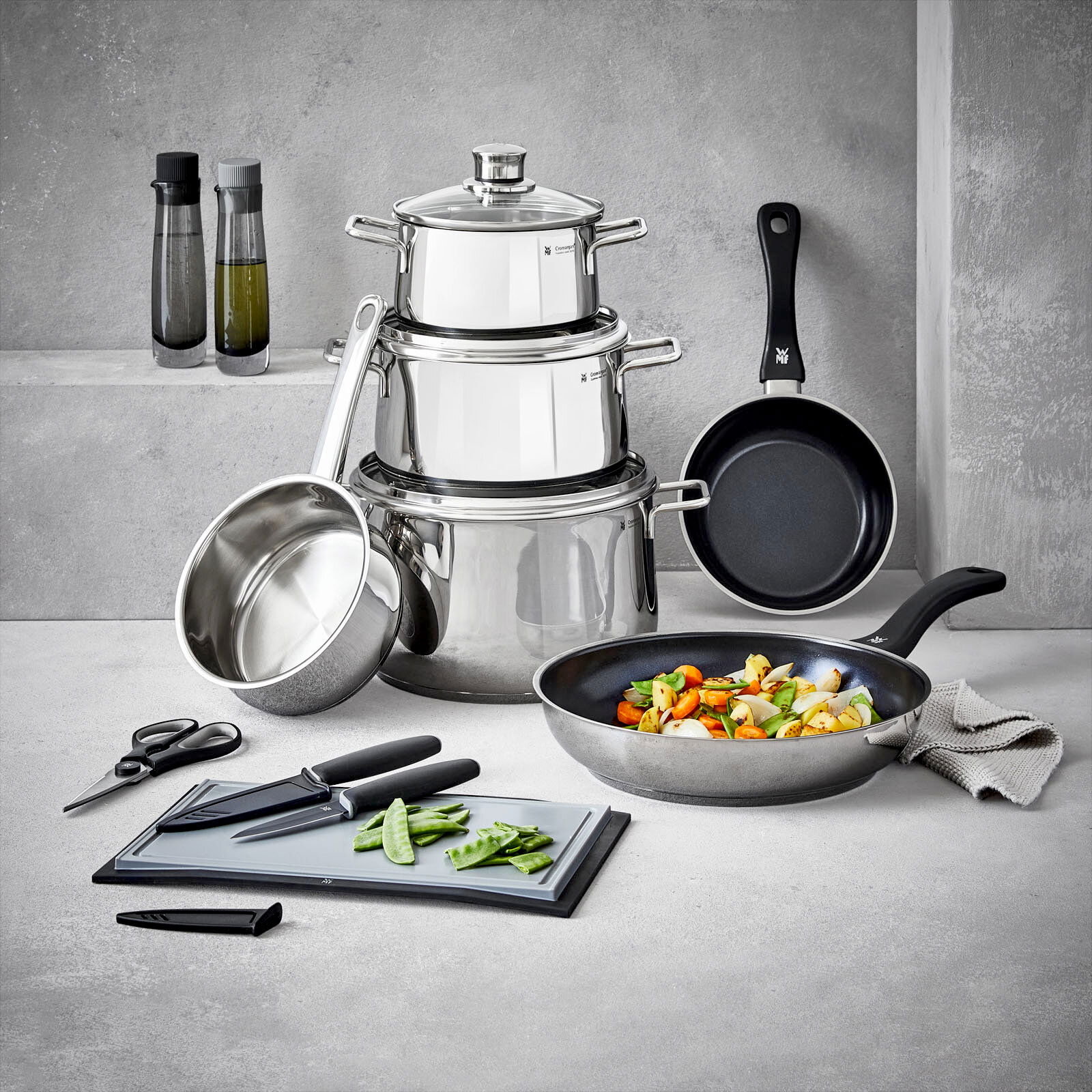 Kochtopf-Starter-Set 10-tlg. aus Edelstahl, bestehend aus mehreren Töpfen und Pfannen, mit Küchenutensilien wie Messer und Schneidebrett, in einer modernen Küchenumgebung von vorne fotografiert.