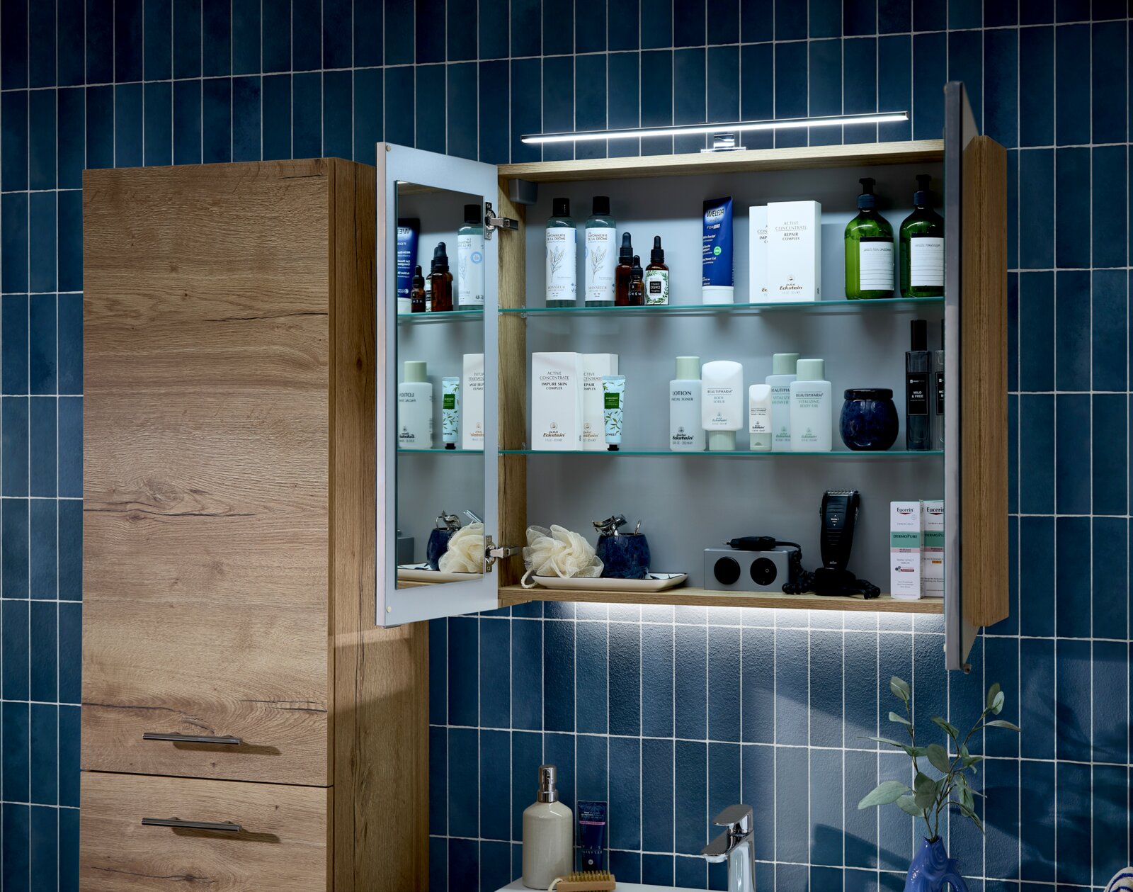 Offener Badezimmerschrank mit Holzoptik und integrierter Spiegel-Tür, Glasablagen und LED-Beleuchtung über dem Waschtisch vor blauer Fliesenwand, frontal aus leicht schräger Perspektive aufgenommen.