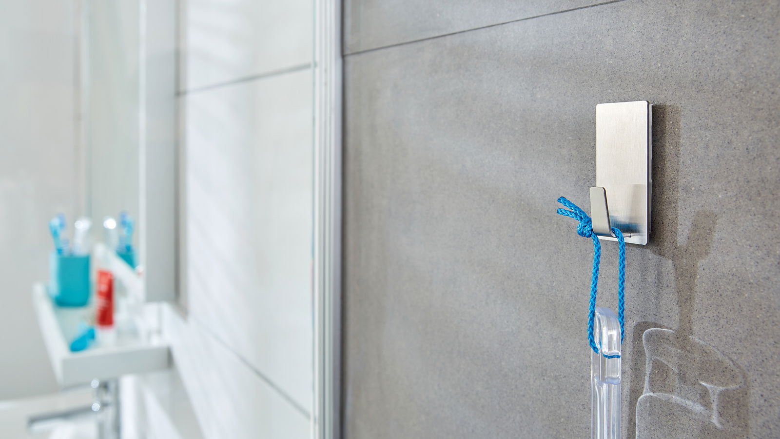 Tesa Powerstrips WATERPROOF Nahaufnahme eines tesa Powerstrips Waterproof Hakens an einer Badezimmerwand, an dem ein blauer Faden mit einem transparenten Gegenstand hängt.