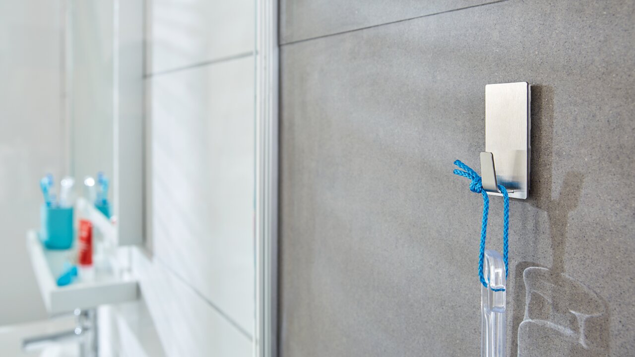 Nahaufnahme eines tesa Powerstrips Waterproof Hakens an einer Badezimmerwand, an dem ein blauer Faden mit einem transparenten Gegenstand hängt.