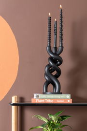 Schwarzer, gedrehter Kerzenhalter mit drei brennenden Kerzen, frontal auf einem Regal mit Büchern und einer Pflanze im Vordergrund.