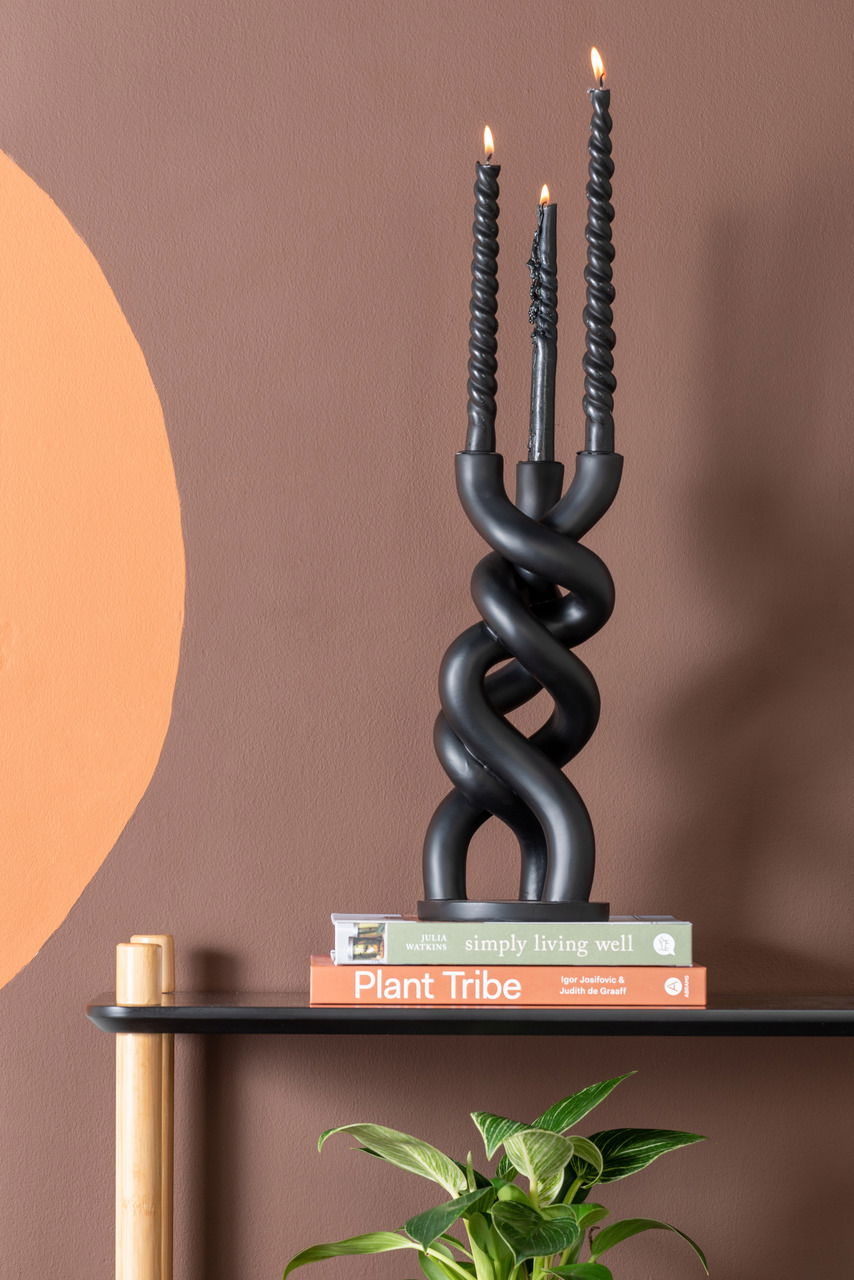 Schwarzer, gedrehter Kerzenhalter mit drei brennenden Kerzen, frontal auf einem Regal mit Büchern und einer Pflanze im Vordergrund.
