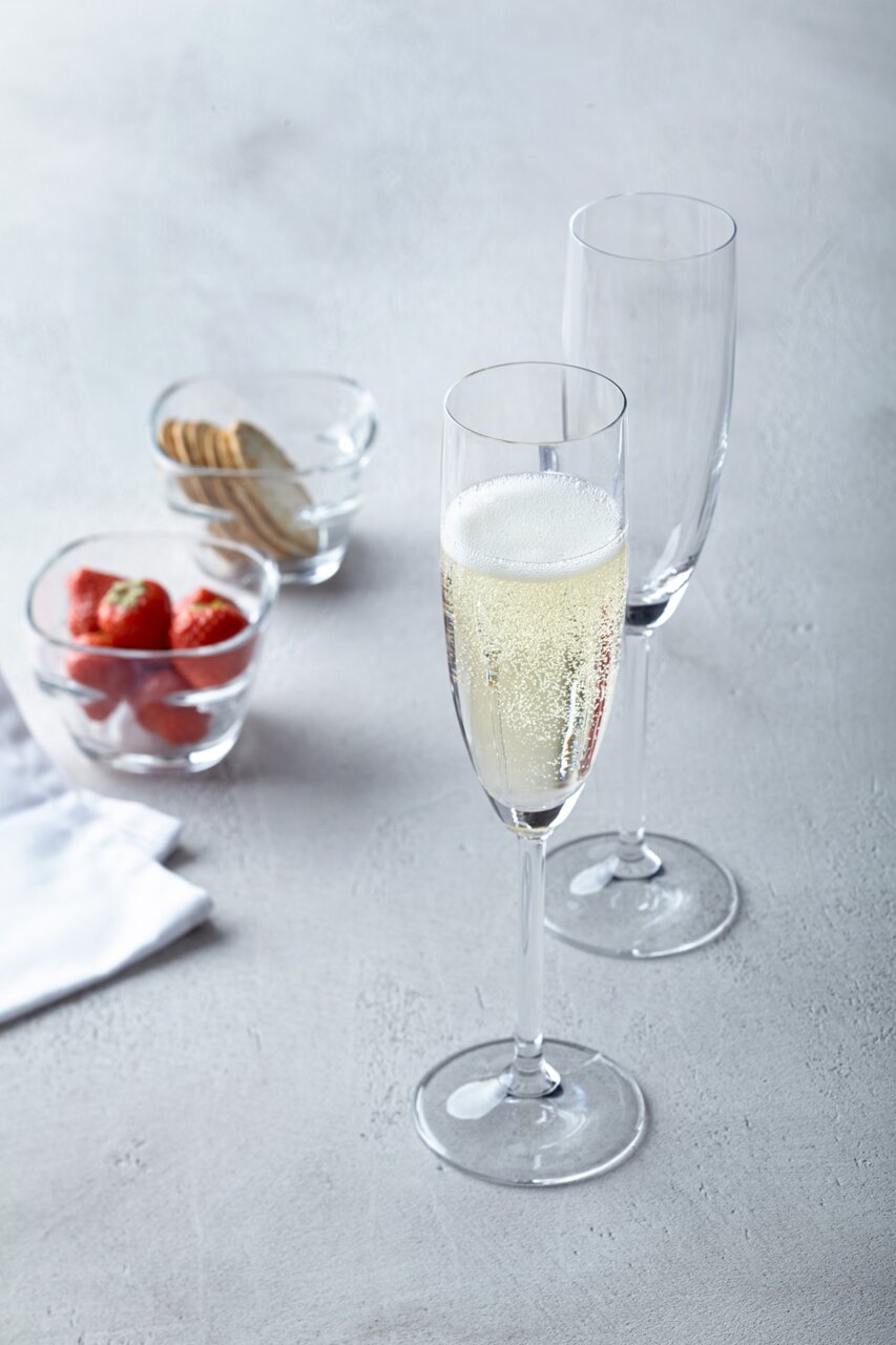 Twee elegante champagneglazen, waarvan één gevuld met bruisende champagne, op een tafel met aardbeien en koekjes op de achtergrond, gefotografeerd vanuit een licht verhoogd perspectief.