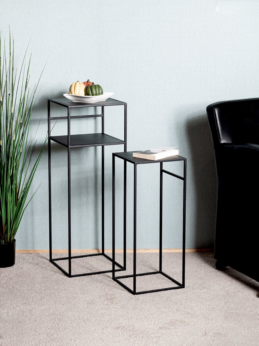 Zwei schwarze, minimalistische Beistelltische aus Metall in unterschiedlichen Höhen neben einem schwarzen Sofa, schräg von vorn in Innenraum-Setting fotografiert; auf dem hohen Tisch steht eine Schale mit Zierkürbissen, auf dem niedrigen liegt ein Buch.