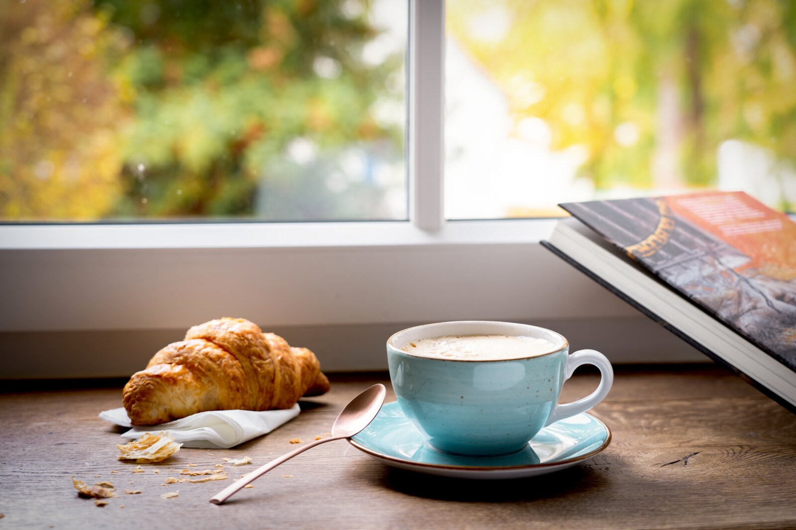 Hellblaue Kaffeetasse mit Untertasse und Milchschaum auf einem Holztisch am Fenster, daneben Croissant auf Serviette, Löffel und ein aufgeschlagenes Buch Perspektive: leicht seitliche Tischhöhe (Eye-Level).
