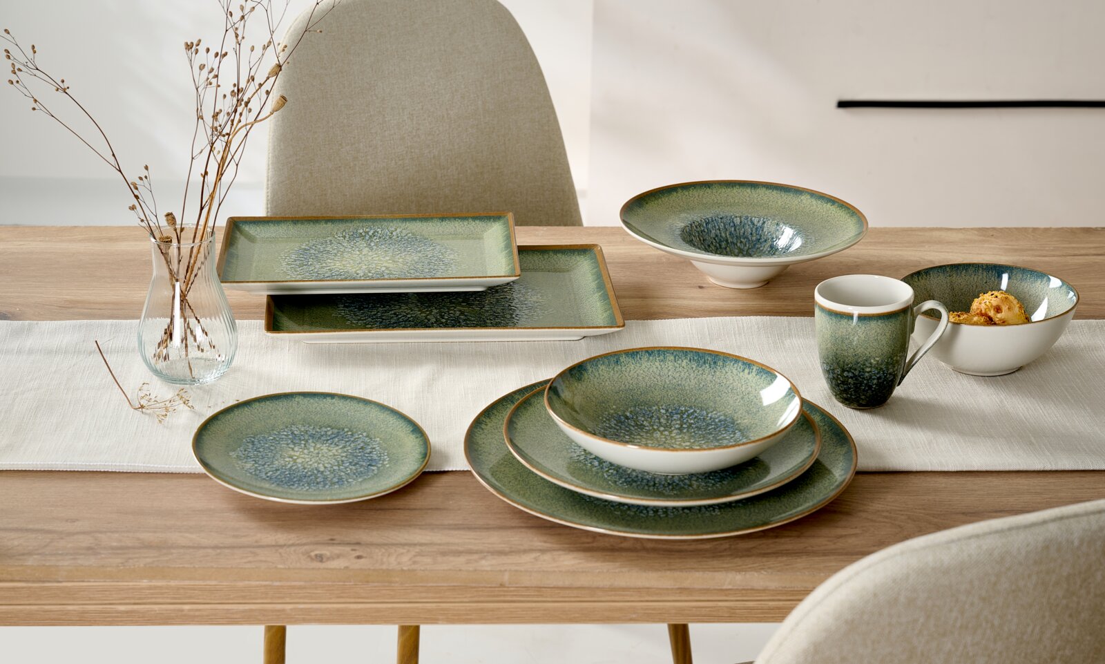 Ritzenhoff & Breker Serviesbord VERDE Schuin van voren gefotografeerd groen-blauw reactief geglazuurd servies met ronde en rechthoekige borden, kommen en mok op een gedekte houten eettafel.