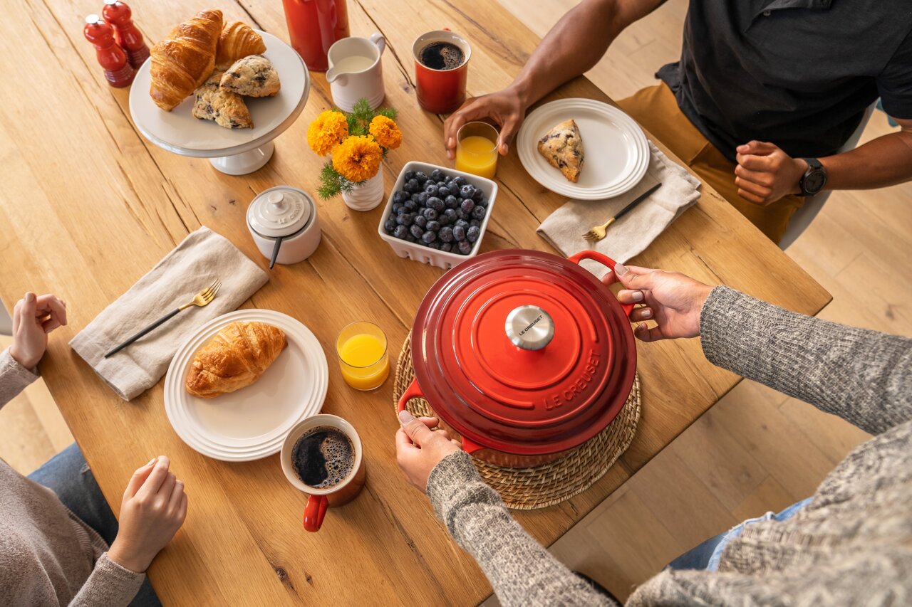 Runder roter Bräter auf einem Esstisch, von oben fotografiert, umgeben von Frühstücksartikeln wie Croissants, Saft und Kaffee.