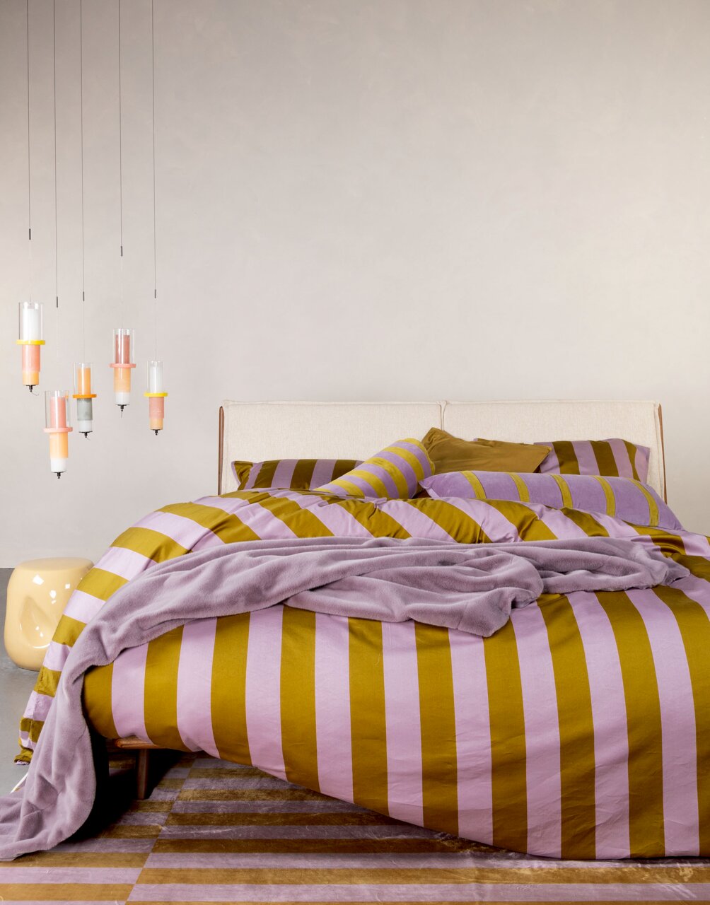 Bett mit gestreifter Bettwäsche in Lila und Senfgelb, dekoriert mit passenden Kissen und einer Decke, aus der Perspektive von vorne fotografiert.