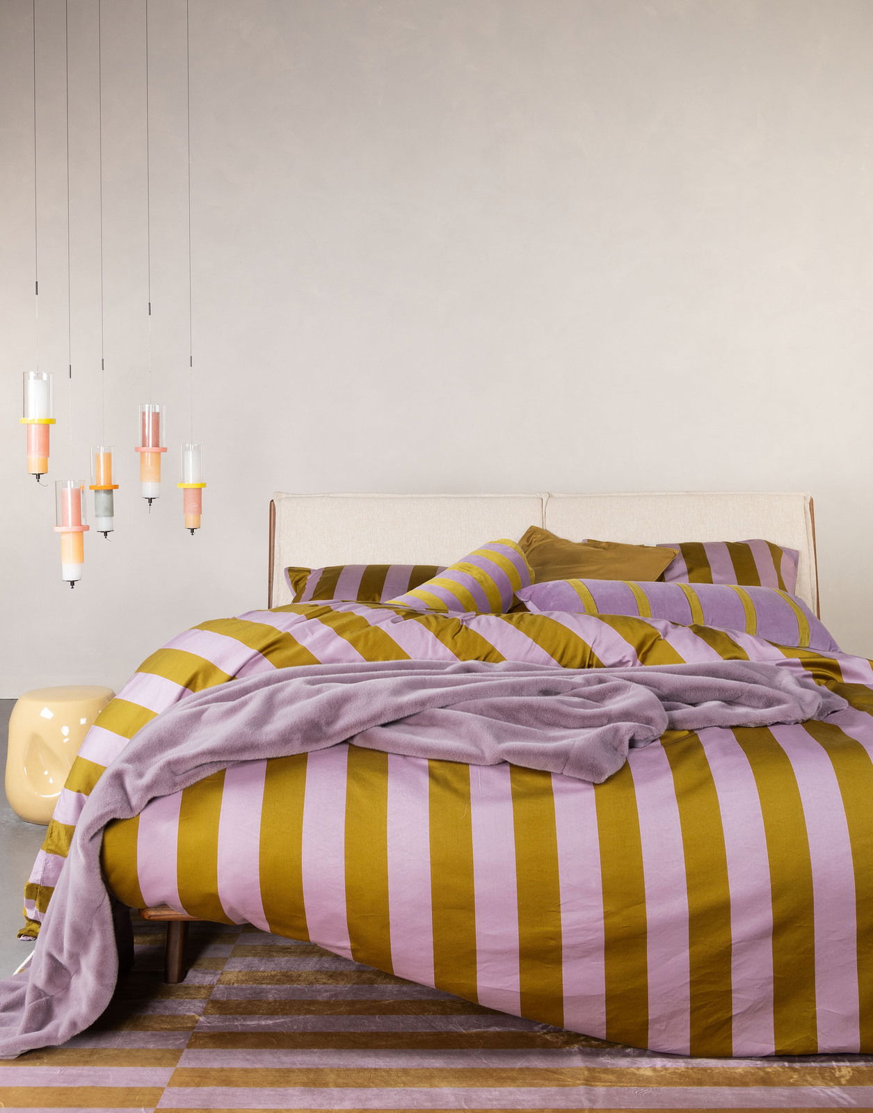 Bett mit gestreifter Bettwäsche in Lila und Senfgelb, dekoriert mit passenden Kissen und einer Decke, aus der Perspektive von vorne fotografiert.