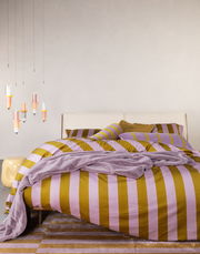 Bett mit gestreifter Bettwäsche in Lila und Senfgelb, dekoriert mit passenden Kissen und einer Decke, aus der Perspektive von vorne fotografiert.