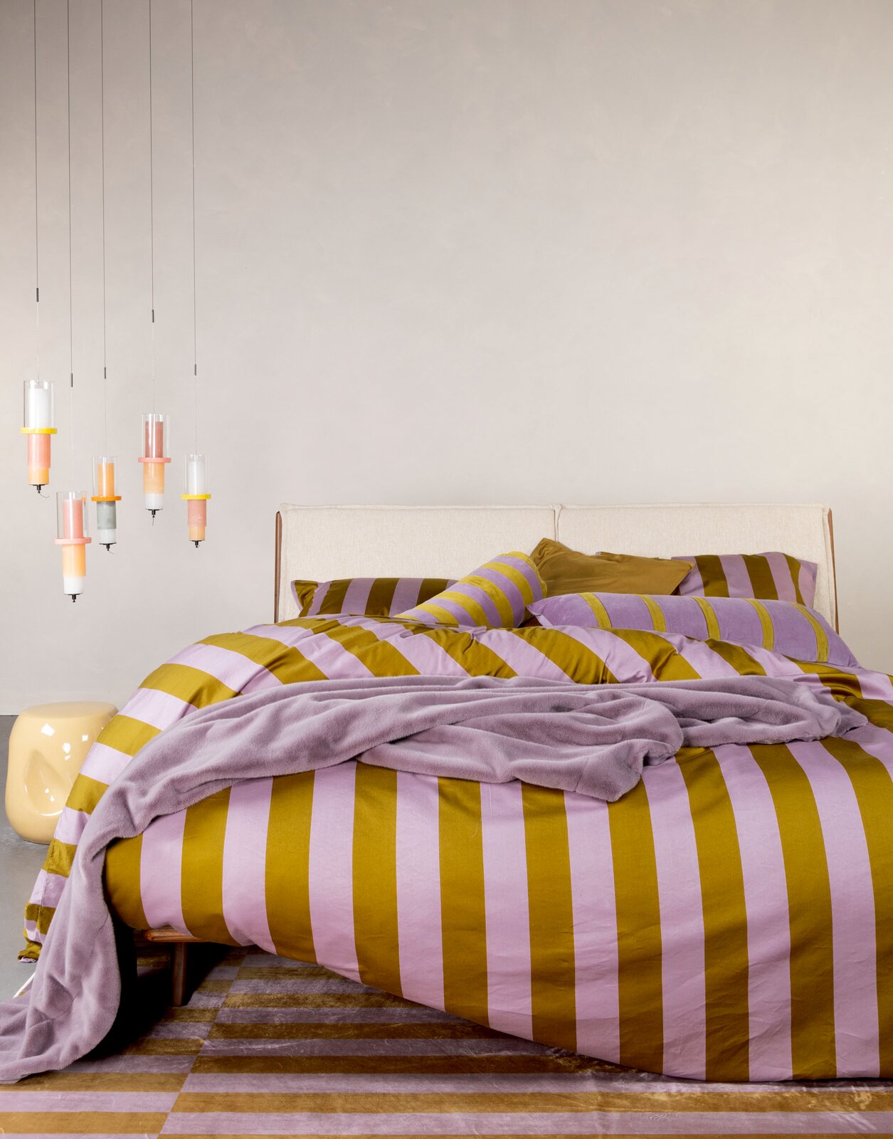Bett mit gestreifter Bettwäsche in Lila und Senfgelb, dekoriert mit passenden Kissen und einer Decke, aus der Perspektive von vorne fotografiert.