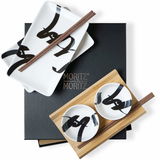 Sushi Set 10-teilig mit schwarzer Schrift, bestehend aus rechteckigen und runden Tellern sowie Essstäbchen, auf einer schwarzen und hölzernen Unterlage von oben betrachtet.