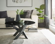 Moderner Couchtisch mit schwarzer Glasplatte und gekreuztem Metallgestell, seitliche Perspektive im Wohnzimmer mit Sofa und Pflanzen.