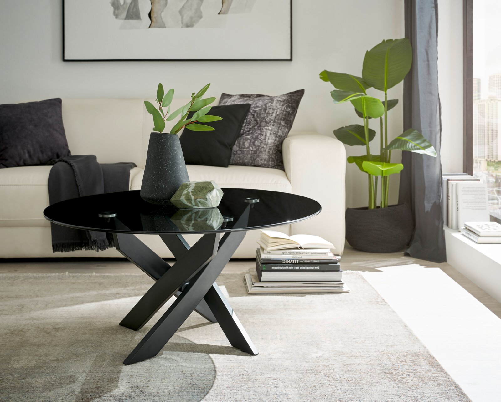 Moderner Couchtisch mit schwarzer Glasplatte und gekreuztem Metallgestell, seitliche Perspektive im Wohnzimmer mit Sofa und Pflanzen.
