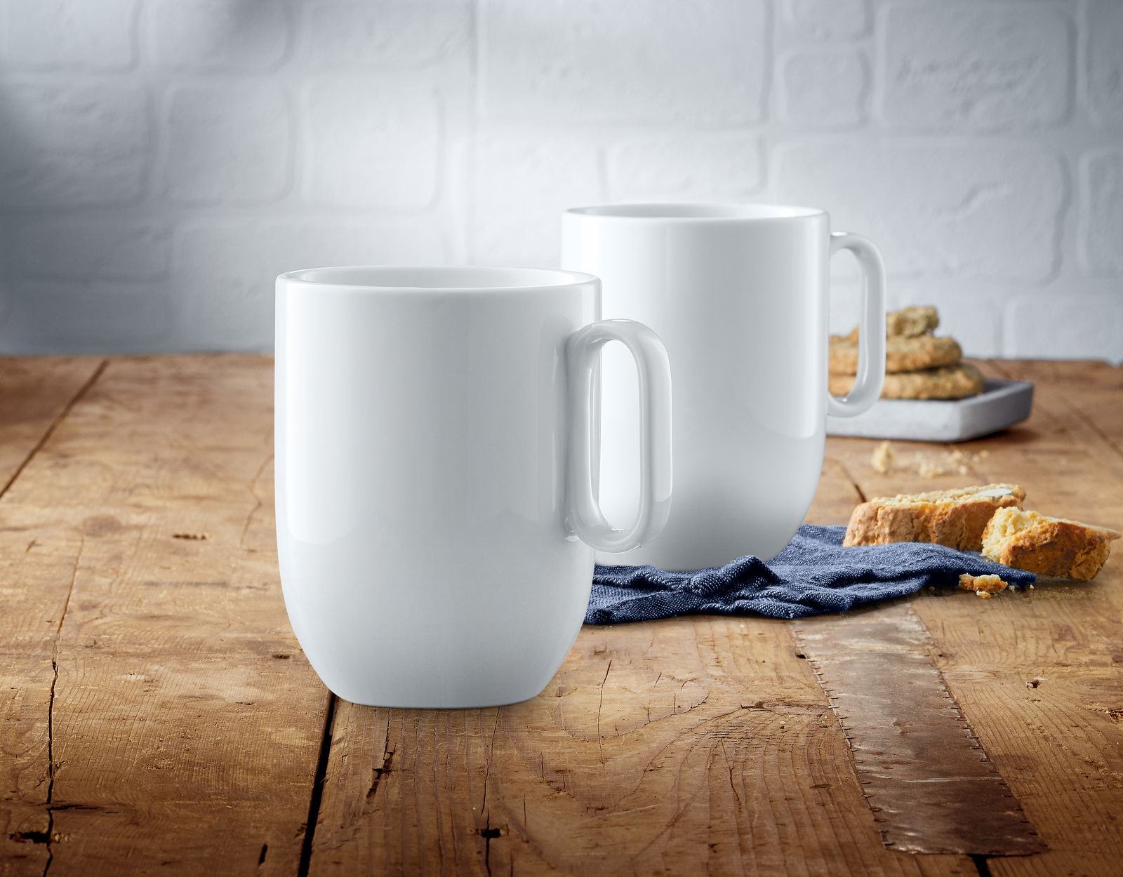 Zwei weiße Kaffeetassen aus dem WMF Barista Kaffeetassen-Set auf einem Holztisch, im Vordergrund eine Tasse, im Hintergrund eine weitere Tasse und Gebäck.