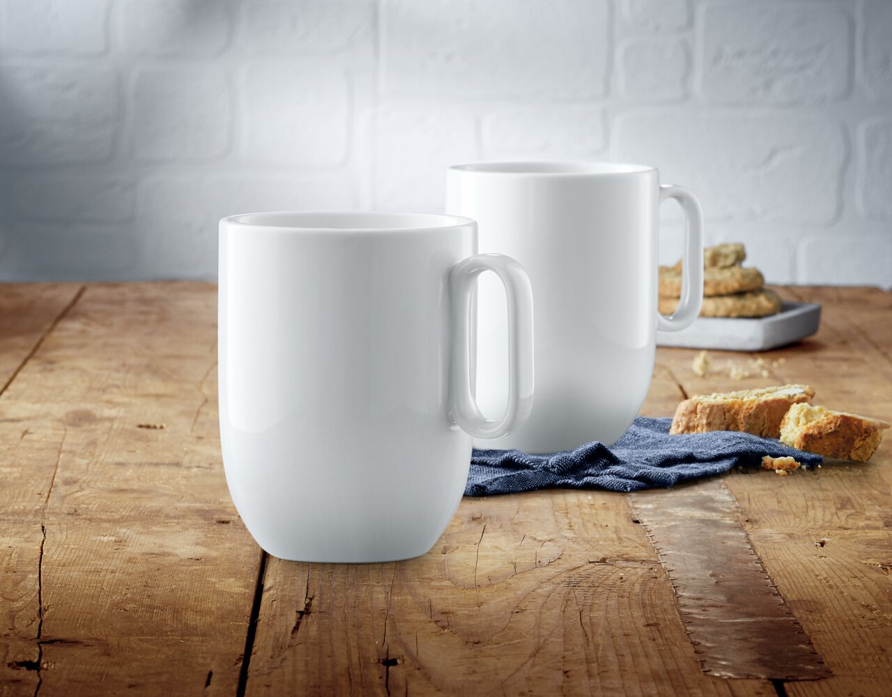 Zwei weiße Kaffeetassen aus dem WMF Barista Kaffeetassen-Set auf einem Holztisch, im Vordergrund eine Tasse, im Hintergrund eine weitere Tasse und Gebäck.