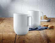 Zwei weiße Kaffeetassen aus dem WMF Barista Kaffeetassen-Set auf einem Holztisch, im Vordergrund eine Tasse, im Hintergrund eine weitere Tasse und Gebäck.