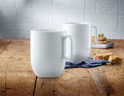 Zwei weiße Kaffeetassen aus dem WMF Barista Kaffeetassen-Set auf einem Holztisch, im Vordergrund eine Tasse, im Hintergrund eine weitere Tasse und Gebäck.