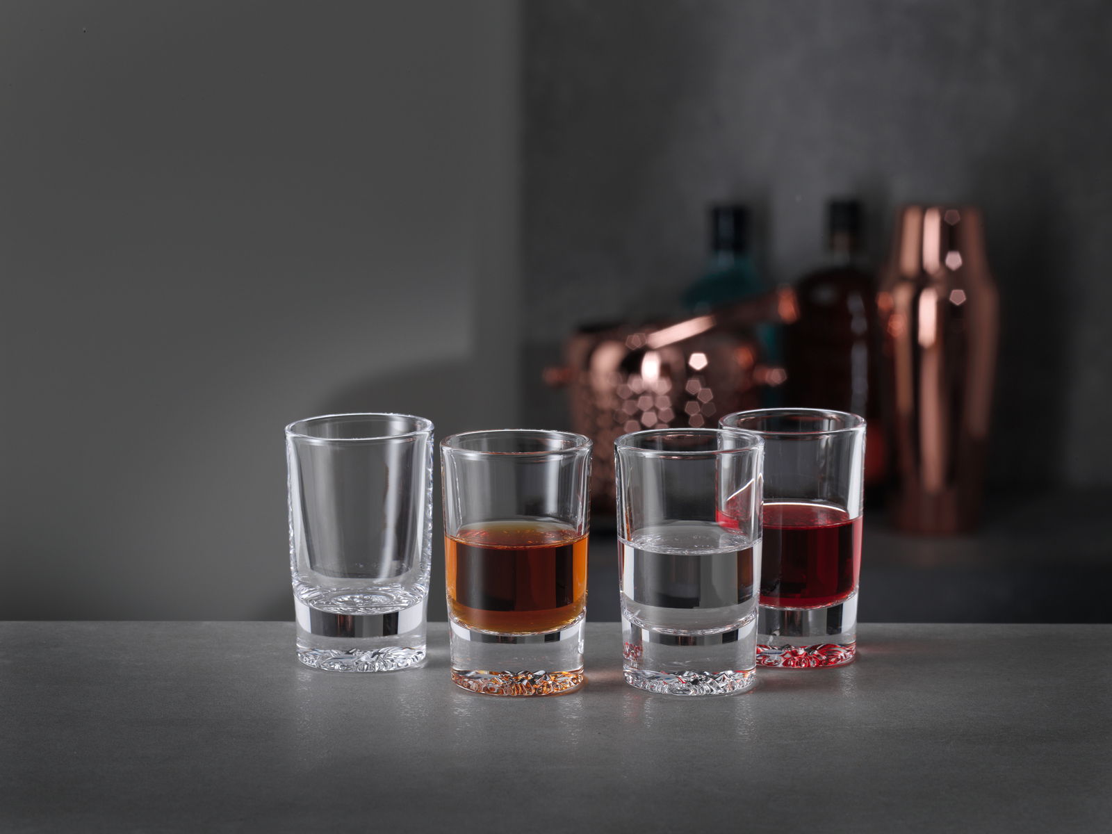 Spiegelau Shot Gläser 4er Set LOUNGE Vier Schnapsgläser in einer Reihe, gefüllt mit verschiedenen Flüssigkeiten, auf einer grauen Oberfläche vor einem unscharfen Hintergrund mit Barzubehör. Perspektive von vorne.