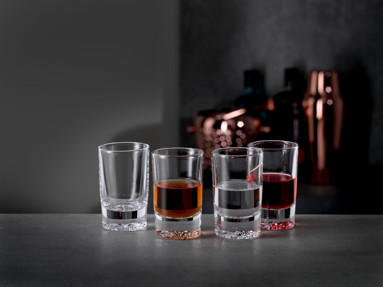 Spiegelau Shot Gläser 4er Set LOUNGE Vier Schnapsgläser in einer Reihe, gefüllt mit verschiedenen Flüssigkeiten, auf einer grauen Oberfläche vor einem unscharfen Hintergrund mit Barzubehör. Perspektive von vorne.
