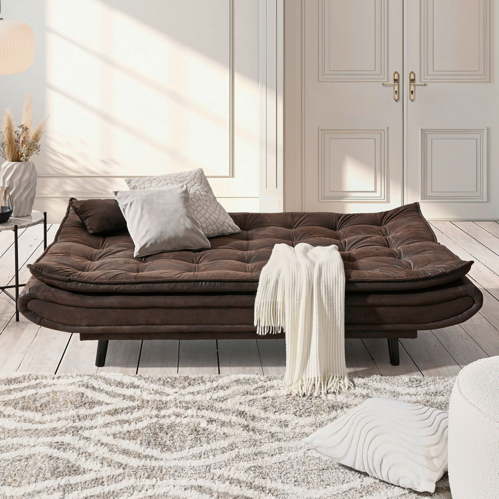 Braunes Funktionssofa in einem hellen Raum, frontal betrachtet, dekoriert mit Kissen und einer weißen Decke.