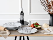 Weißer Pizzateller mit grauem Schriftzug „PIZZA LOVE AT FIRST BITE“ auf rustikalem Holztisch, im Vordergrund frontal aus leicht erhöhter Perspektive; daneben gestapelte Teller, Weinflasche mit Glas, Tomaten, Basilikum und Pizzateig.