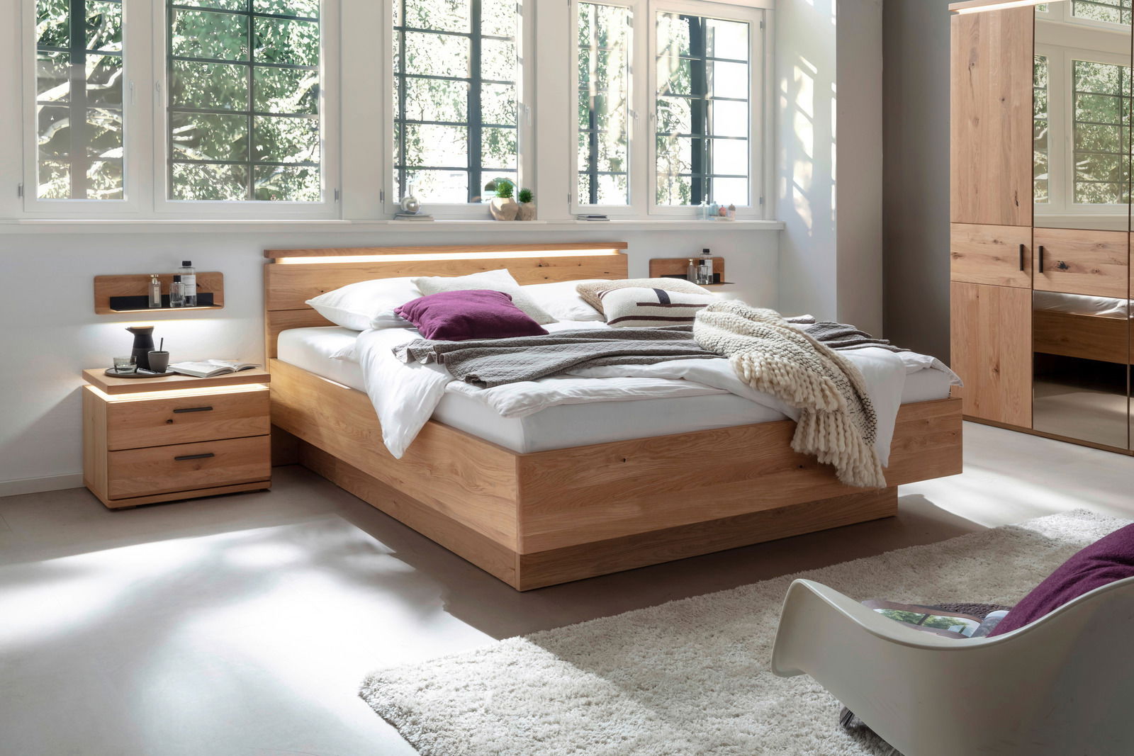 Linea Natura Bett BARCELONA Modernes Schlafzimmer mit einem großen Holzbett, das von der Seite zu sehen ist. Das Bett ist mit weißen und lila Kissen sowie einer gestrickten Decke dekoriert. Neben dem Bett steht ein Nachttisch mit Dekorationen. Im Hintergrund sind große Fenster und e