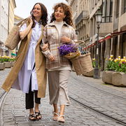 Zwei Frauen spazieren lächelnd auf einer gepflasterten Straße. Eine Frau trägt eine beige Tasche, die andere einen beigen Korb mit lila Blumen. Perspektive: Frontalansicht.