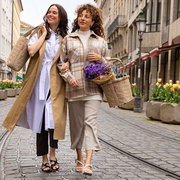 Zwei Frauen spazieren lächelnd auf einer gepflasterten Straße. Eine Frau trägt eine beige Tasche, die andere einen beigen Korb mit lila Blumen. Perspektive: Frontalansicht.