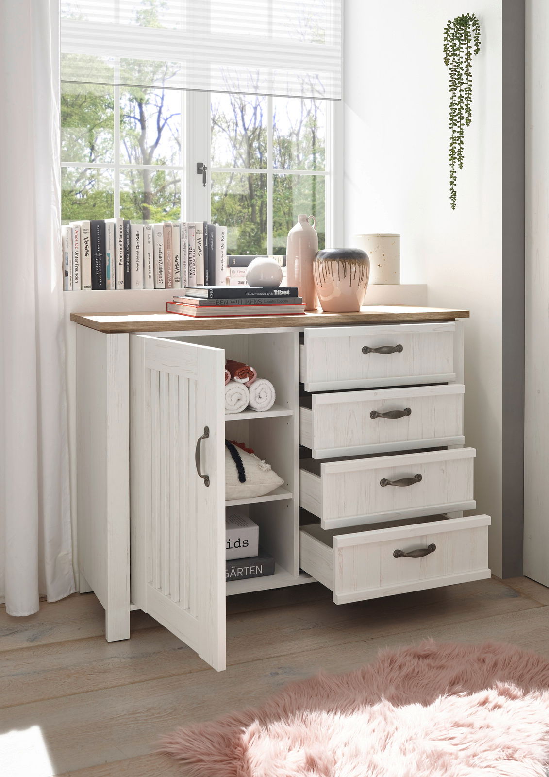 Weiße Kommode mit offenen Schubladen und Tür, dekoriert mit Büchern und Vasen, in einem hellen Raum mit Blick aus dem Fenster.
