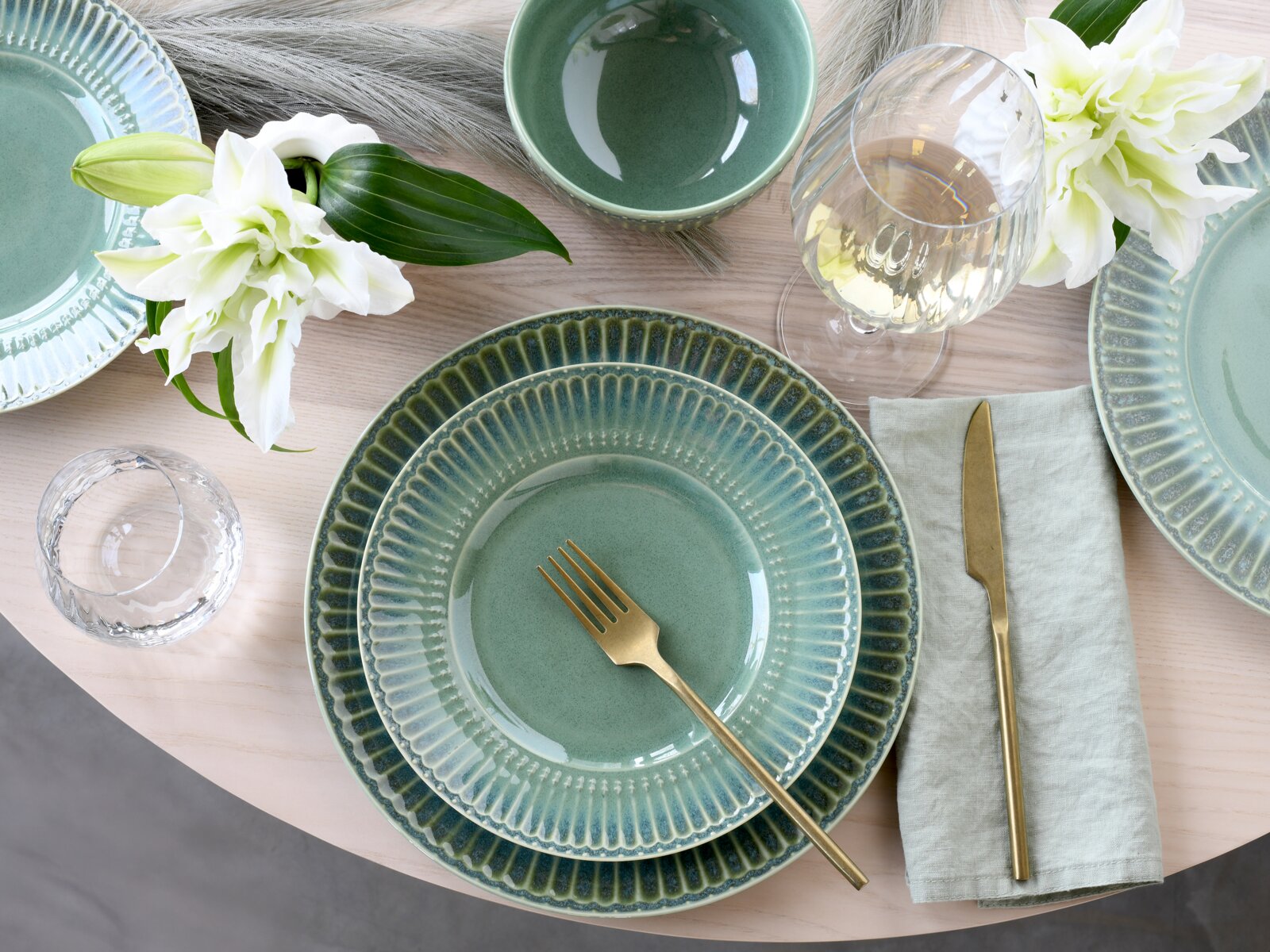 Draufsicht auf eine elegant gedeckte Tafel mit mintgrünem Keramikgeschirr mit geriffeltem Rand, goldfarbenem Besteck, Weinglas und Wasserbecher sowie weißen Lilien auf hellem Holztisch.