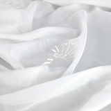 Nahaufnahme eines weißen Vorhangs mit dezentem Blattmuster, ausgebreitet in weicher, fließender Perspektive.