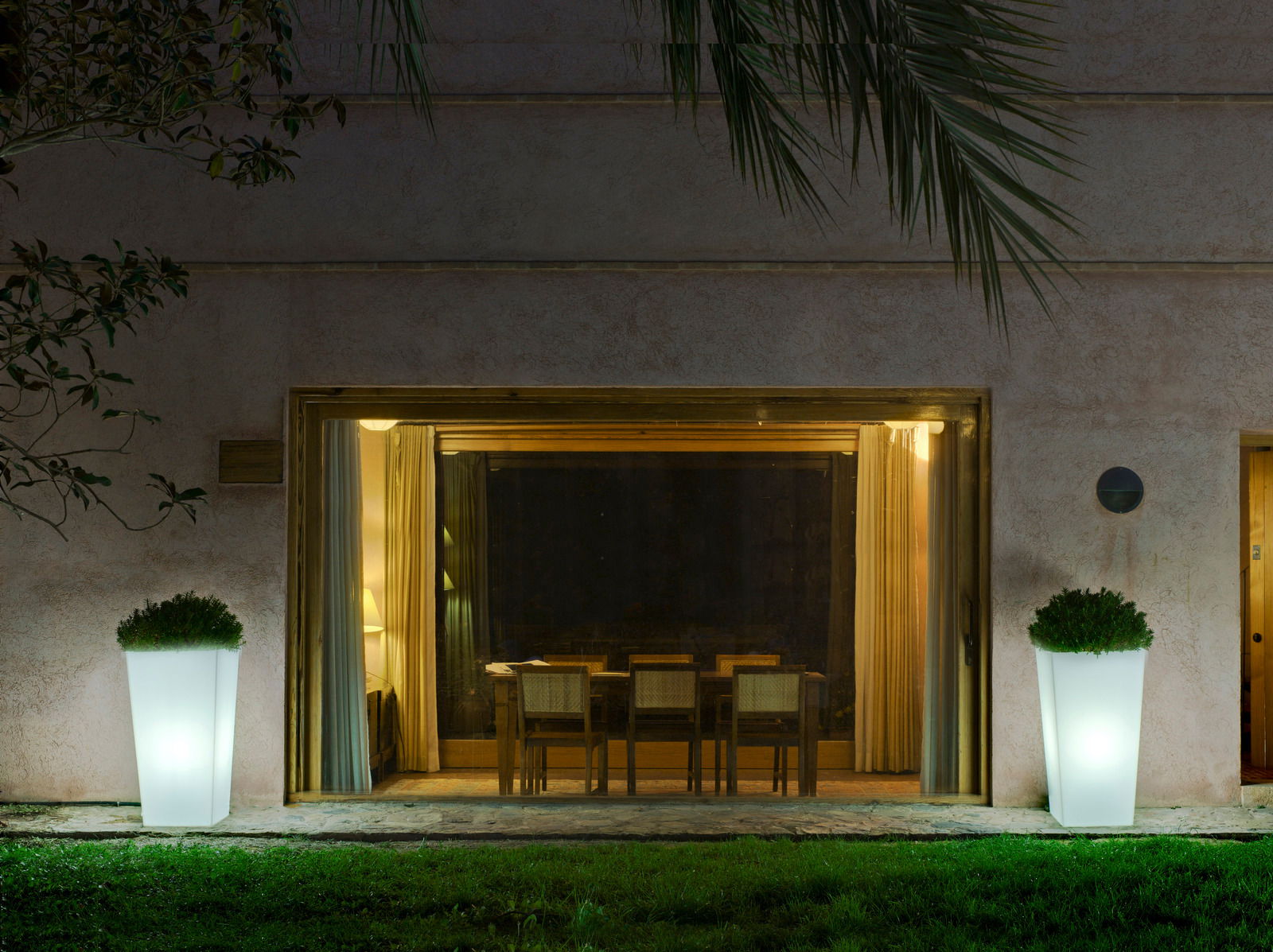 Zwei beleuchtete Blumentöpfe Melisa 50 vor einem großen Fenster, nachts im Garten platziert, mit Blick auf das Haus.